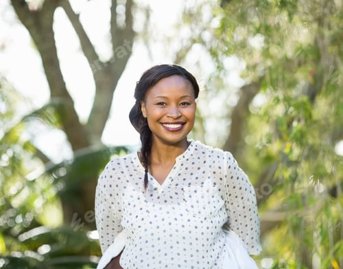 Preview: Mid-adult African American woman smiling while standing in sunlit garden, with polka-dot blouse