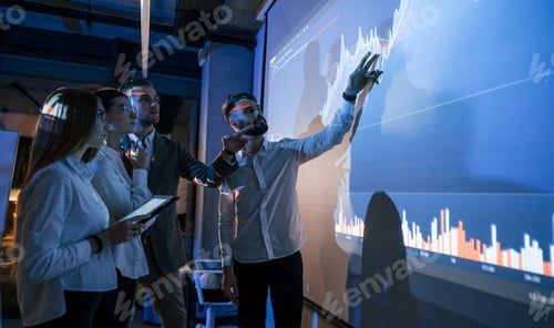 Preview: Holding tablet. Male leader talking to employees, showing the plan on the projector in office