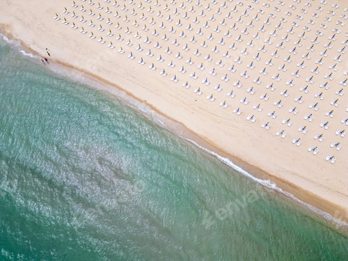 Visualização: Uma praia de areia se estende à beira-mar, adornada com espreguiçadeiras vazias. Vista aérea