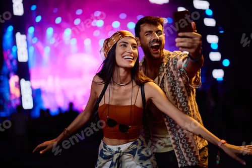 Preview: Cheerful couple using cell phone while taking selfie on summer music festival at night.
