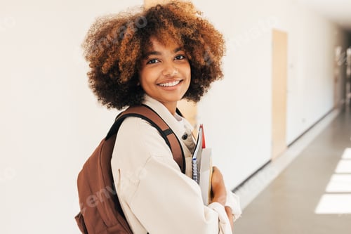 Preview: Smiling girl standing in college corridor holding textbooks and looking at camera