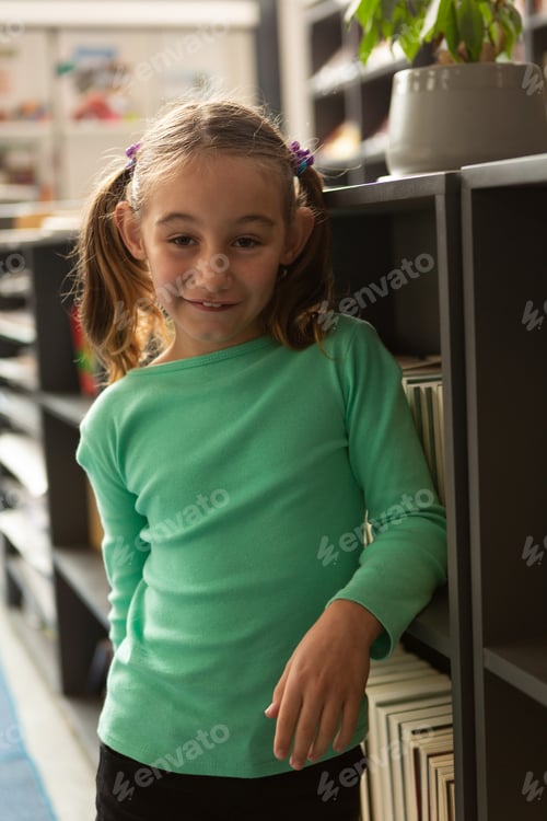 Preview: Front view of smiling Caucasian schoolgirl standing in library at elementary school
