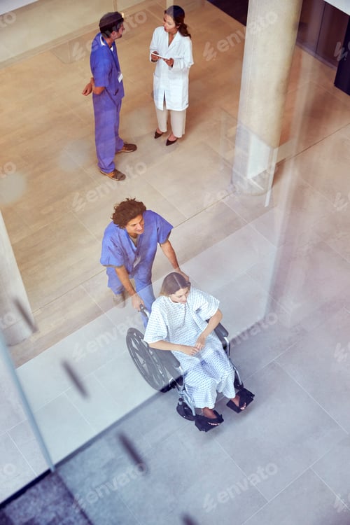 Preview: Overhead Shot Of Nurse Wearing Scrubs Pushing Female Patient In Wheelchair Through Hospital Building