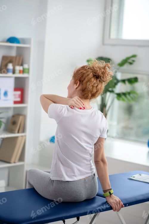 Preview: Red-haired female patient sitting on couch and massaging her painful neck