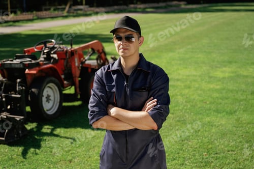 Preview: with arms crossed. Man is with utility tractor with grass cutter and aerator equipment on the field