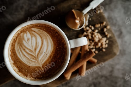 Preview: Cup of coffee with latte art on a wooden board, accompanied by cinnamon sticks, nuts, and caramel