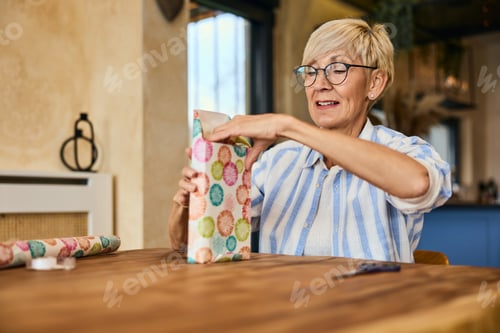 Preview: A senior woman using wrapping paper, packing a gift for someone, sitting at home.