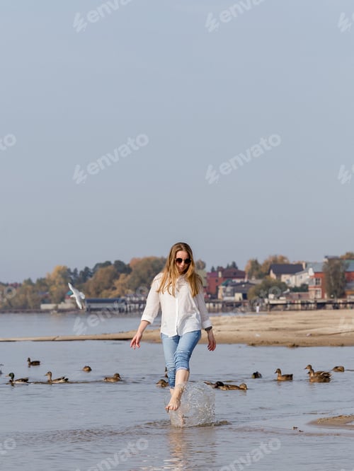 Preview: young woman in a white shirt and jeans walks barefoot along the banks of the river