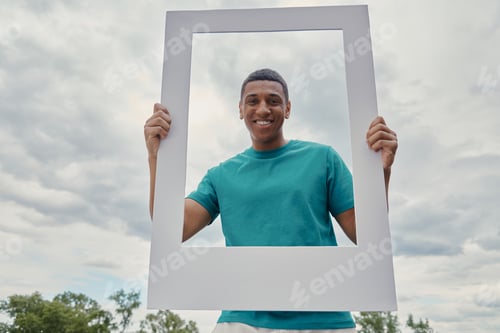 Preview: Happy mixed race man looking through a picture frame and smiling while standing outdoors