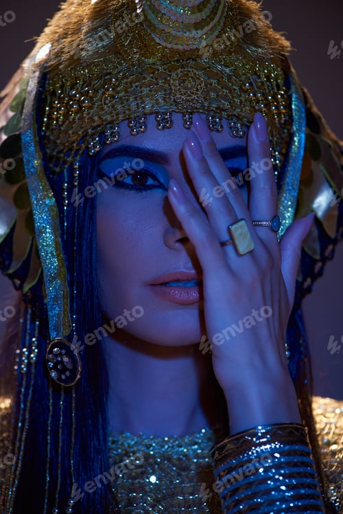 Preview: Portrait of woman in Egyptian headdress covering face and posing on brown background with blue light