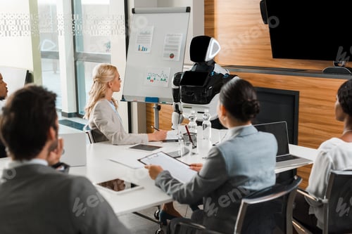 Preview: young multicultural businesspeople sitting in conference hall and looking at robot