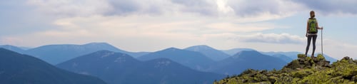 Preview: Back view of young slim backpacker tourist girl with stick standing on rocky top
