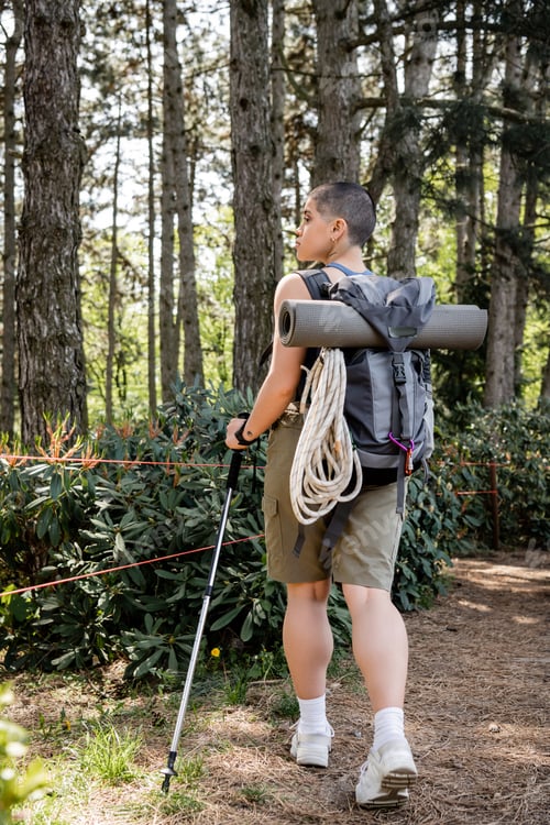 Preview: Side view of young short haired woman traveler with backpack holding trekking pole and hiking