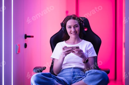 Preview: Young girl playing mobile online game on a smart phone while sitting on gaming chair in colorful