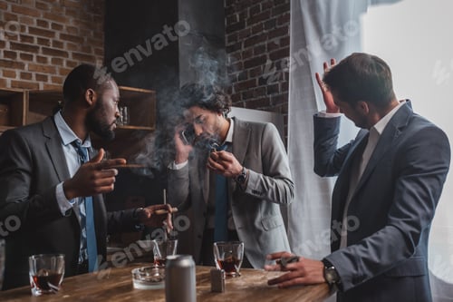 Preview: emotional young businessman smoking cigar and talking by smartphone while friends drinking whiskey