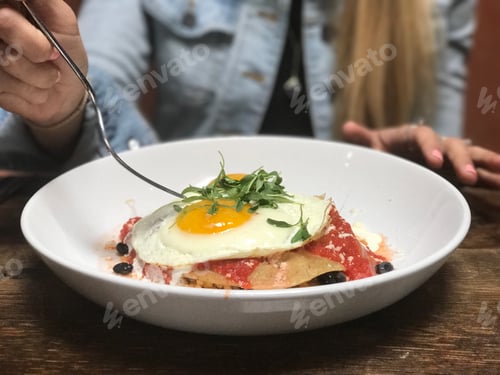 Preview: A woman uses her fork to take her first bite of huevos rancheros with an egg on top for breakfast.
