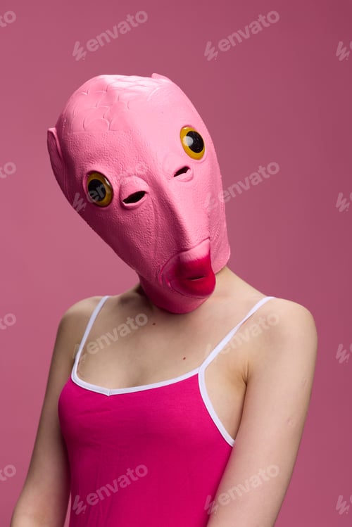 Preview: A woman wearing a pink fish head mask on Halloween stands and looks at the camera against a pink