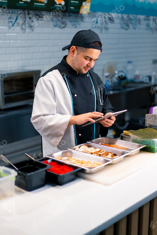 Preview: Thoughtful Sushi Chef with a tablet in his hands near the kitchen table on which products for rolls