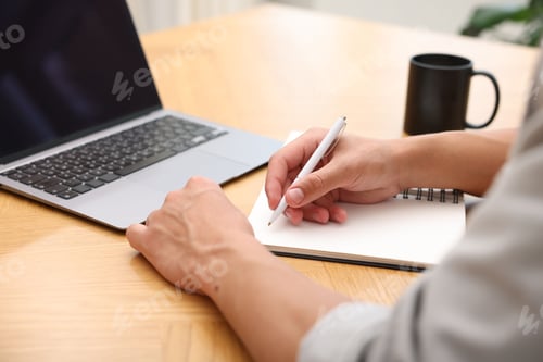 Preview: Copywriter with laptop writing in notebook at wooden table indoors, closeup