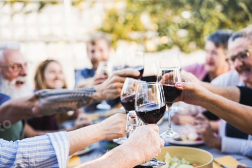 Preview: Happy family cheering with red wine at barbecue dinner outdoor - Focus on front hand holding glass