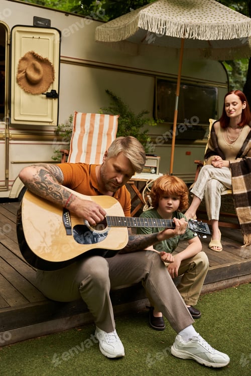 Preview: tattooed man playing acoustic guitar to smiling son near family and trailer home, bonding moments