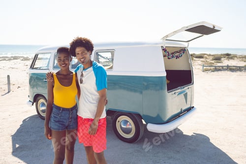 Preview: Front view of diverse couple looking at camera while standing near camper van at beach