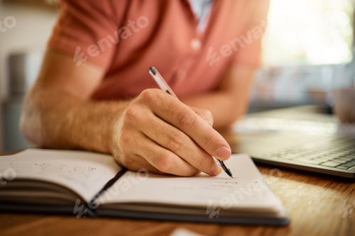 Preview: Man writing in a diary while working from home alone. One businessperson working on a laptop and wr