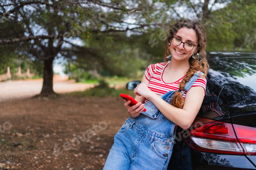 Preview: Young digital nomad woman using smartphone leaning on car in nature