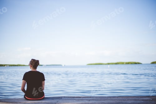 Preview: Female sitting on a wooden pathway near the sea with a blurred background shot from behind