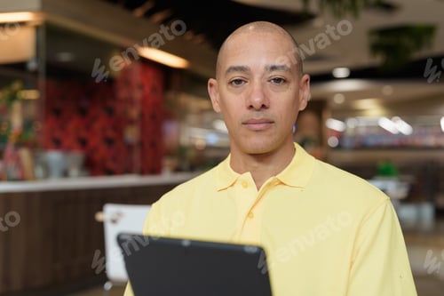 Preview: Middle-aged bald Hispanic man using tablet computer while dining at food court restaurant