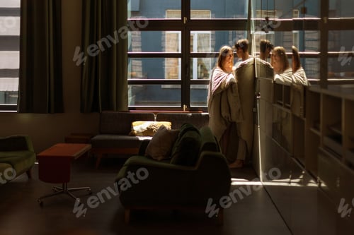 Preview: young lovers at home wrapped in a blanket under a large window