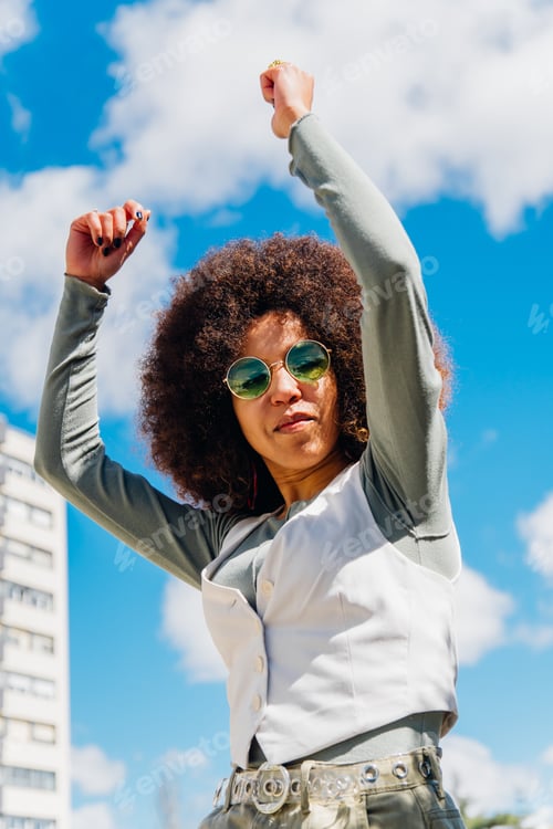 Preview: Young woman with curly hair and sunglasses dancing outdoors in a sunny day with blue sky