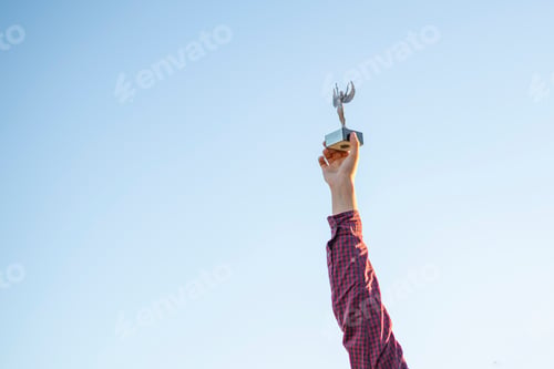 Preview: young person winning a competition, raised hands with a trophy figure against the sky with copy spac