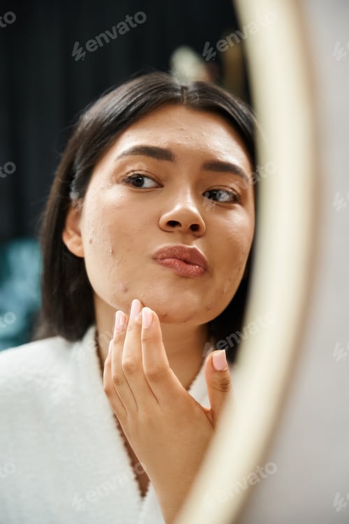 Preview: young asian girl with brunette hair examining her face with acne in bathroom mirror, skin issue