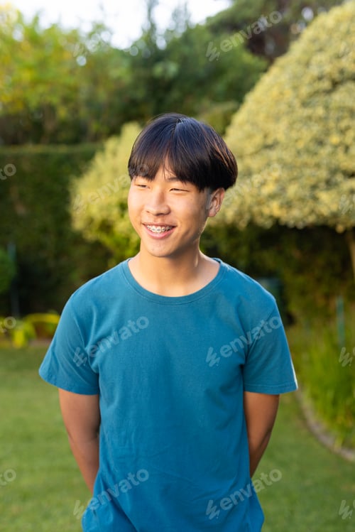 Preview: Smiling young asian teenage boy in blue shirt standing in garden, enjoying outdoor time