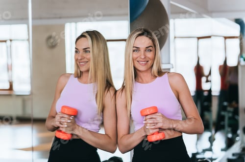 Preview: Smiling woman holding dumbbells in gym, reflection visible in the mirror