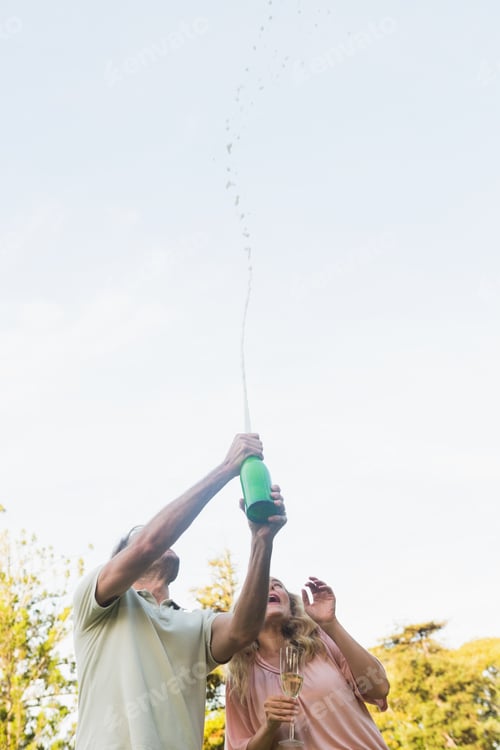 Preview: Handsome man spraying bottle of champagne with blonde partner outside in the sunshine