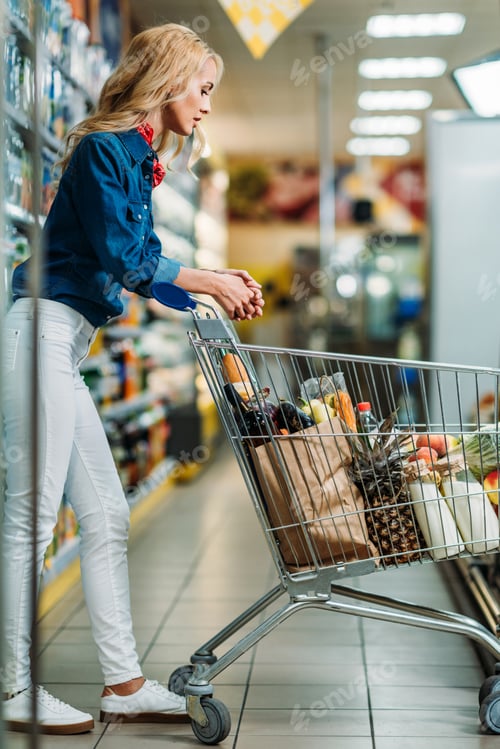 Preview: side view of young woman looking at purchases in shopping cart in supermarket