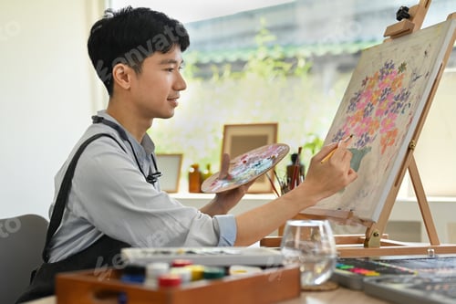 Preview: Image of pleased man artist painting picture with watercolor on canvas in art workshop.