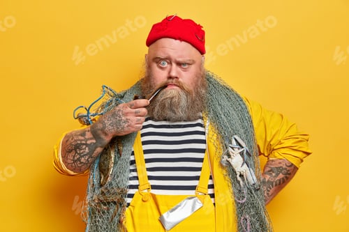 Preview: Close up shot of serious bearded sailor smokes pipe and looks unhappily at camera, poses with fishin
