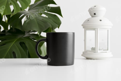Preview: Black mug mockup with a candle holder on a white table and a monstera plant.