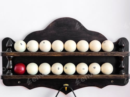 Preview: Billiard balls and triangle for pyramid are on wooden shelf attached to light colored wall.