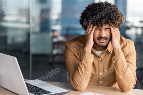Preview: Tired young businessman working inside office at workplace, hispanic man has severe headache stress