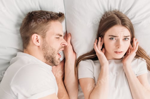 Preview: top view of dissatisfied woman plugging ears with hands and looking at camera while lying in bed