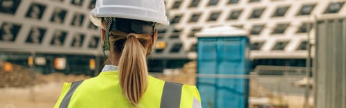 Preview: Back view of female construction worker in protective helmet against on construction background