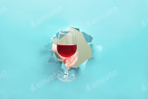 Preview: Hand holds a glass of red wine through a torn hole. Event celebration concept.