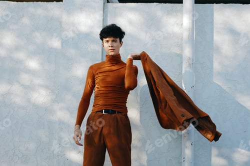 Preview: Young man in a brown turtleneck outfit gracefully holding a jacket against a light textured wall