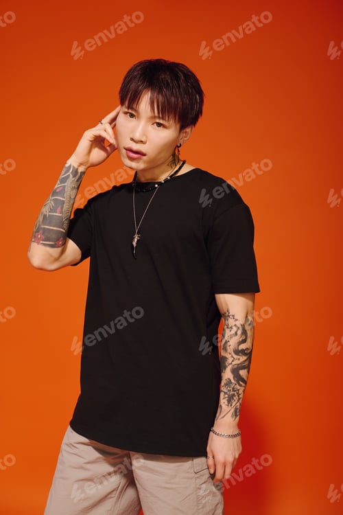 Preview: Stylish young Asian man with tattoos poses confidently against a vibrant orange backdrop