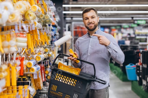 Preview: Man smiling, making DIY purchases, holding a paint roller and basket in a hardware store aisle