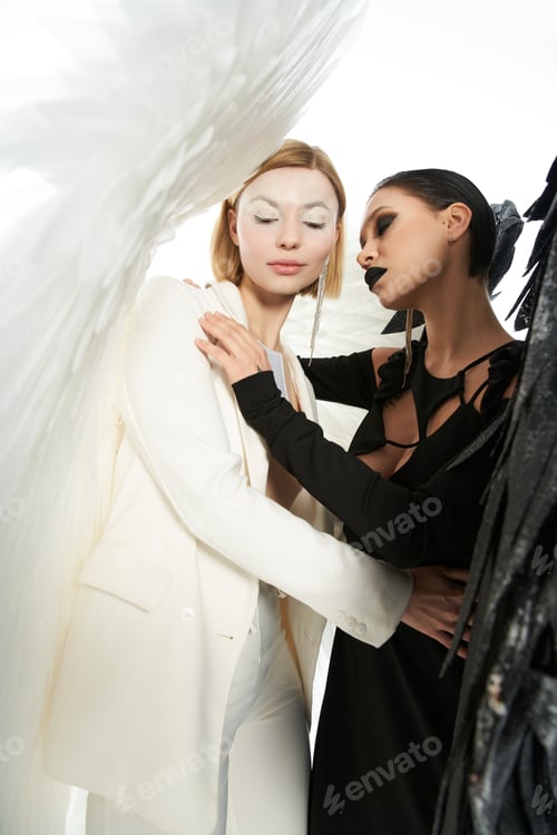 Preview: women in costumes of dark and light winged creatures embracing on white, angel and demon concept